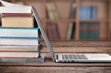 Stack of books next to an open laptop on a wooden table.