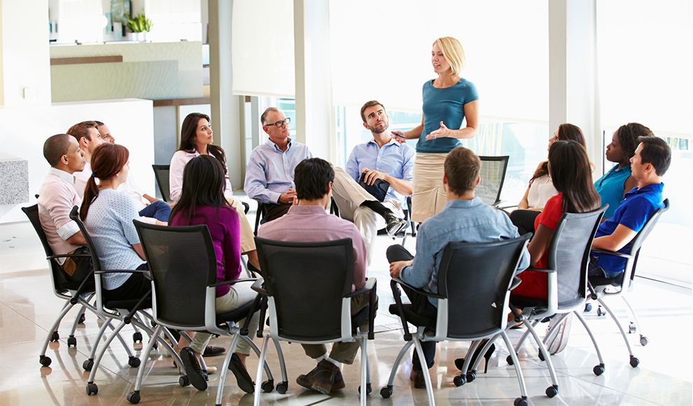Group of people in a meeting, with a woman speaking to them.
