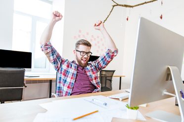 Man celebrating with raised hands while sitting at a computer in an office.