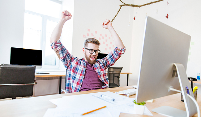 Man celebrating with raised hands while sitting at a computer in an office.