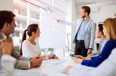 Man giving a presentation to colleagues while showing a chart on a whiteboard.