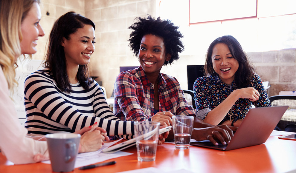 Four smiling women working together on a laptop in an office.
