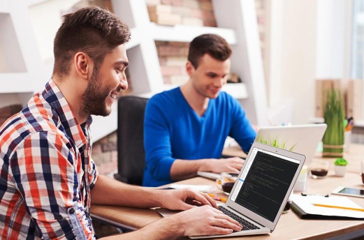 Two male programmers smiling while working on laptops in an office.
