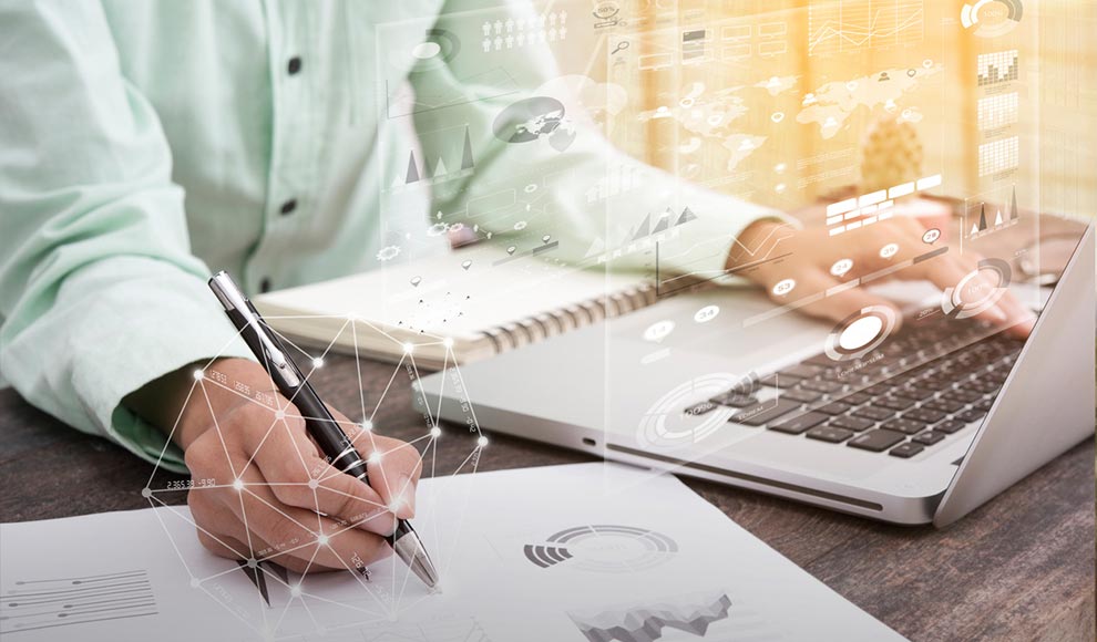 Person writing on paper while working on a laptop with charts.