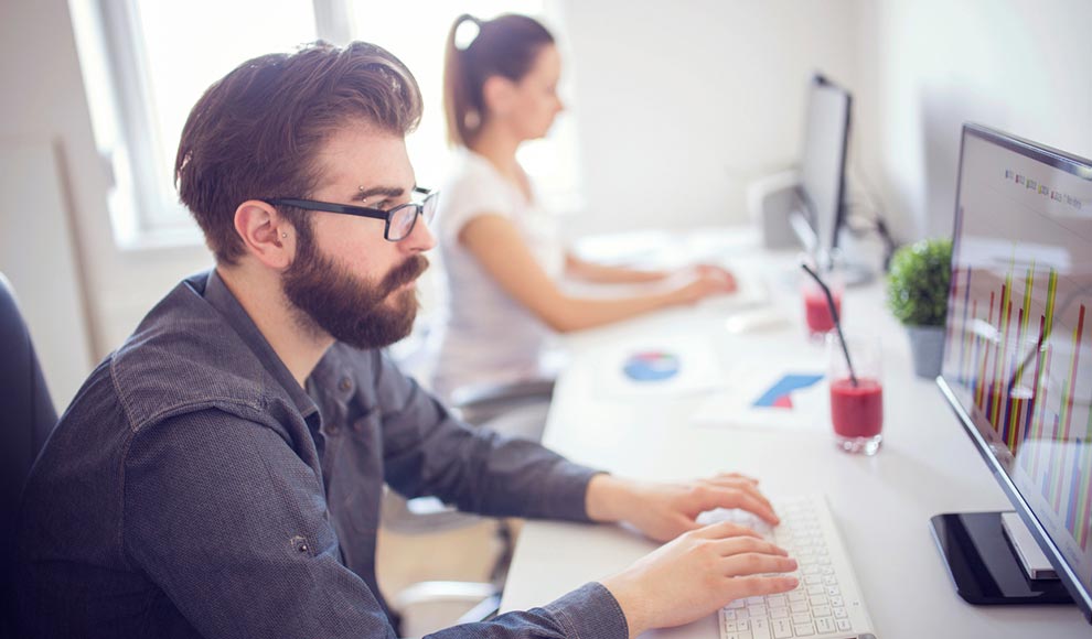 Bearded man with glasses working on a computer in an office with a female colleague.