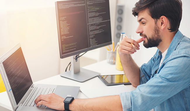 Young programmer working on a laptop and desktop computer, writing code.