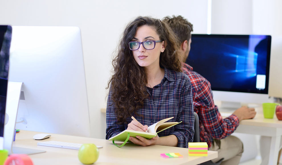 Young woman in a checkered shirt working on a computer and writing in a notebook.