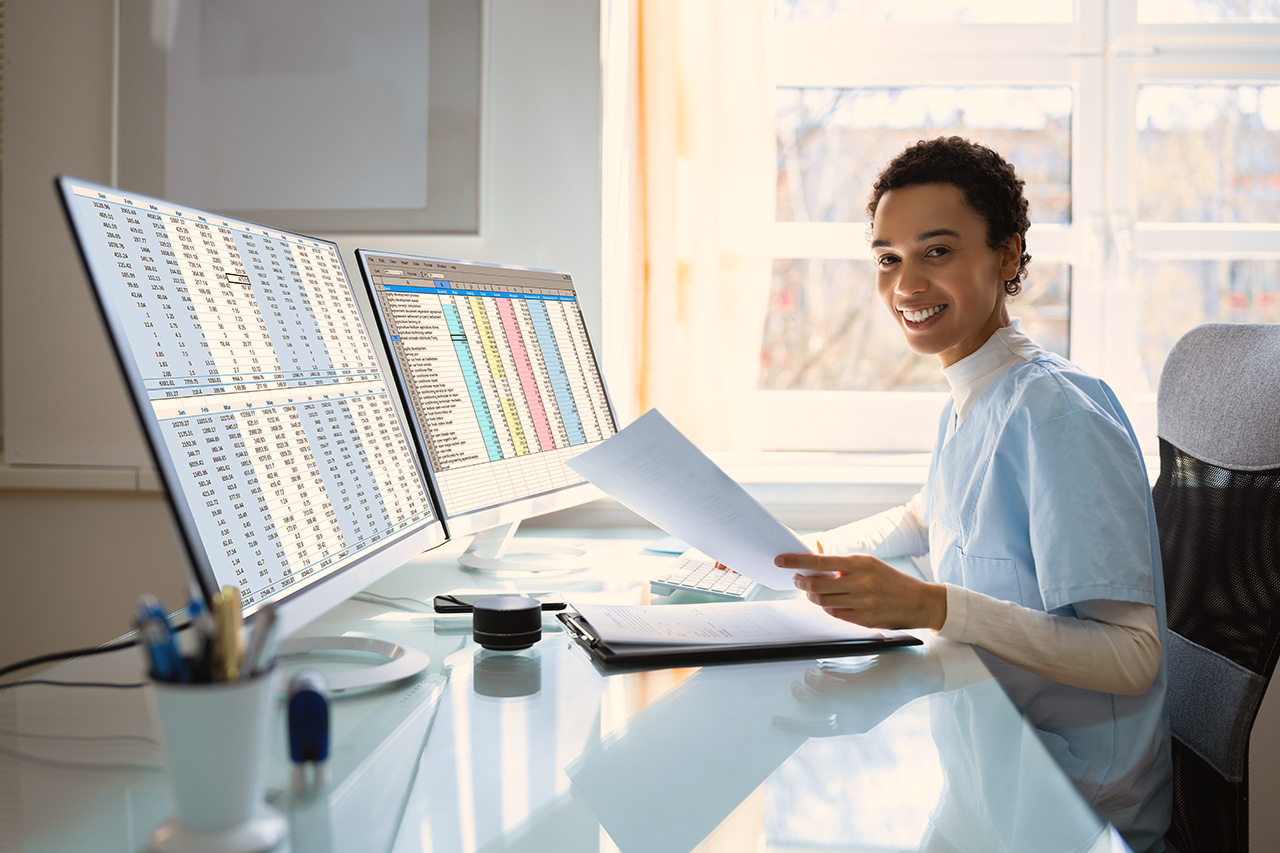 Smiling female doctor sitting at a desk with two monitors showing tables.