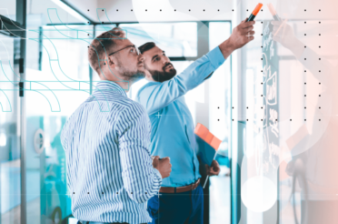 Two businessmen writing on a whiteboard while discussing ideas.