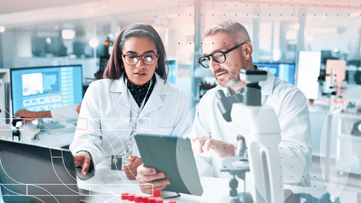 Two scientists discussing data on a tablet in a laboratory while looking at a microscope.