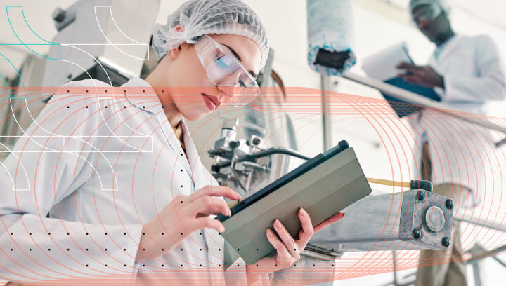 Woman in lab coat and safety glasses using a tablet in a manufacturing environment.